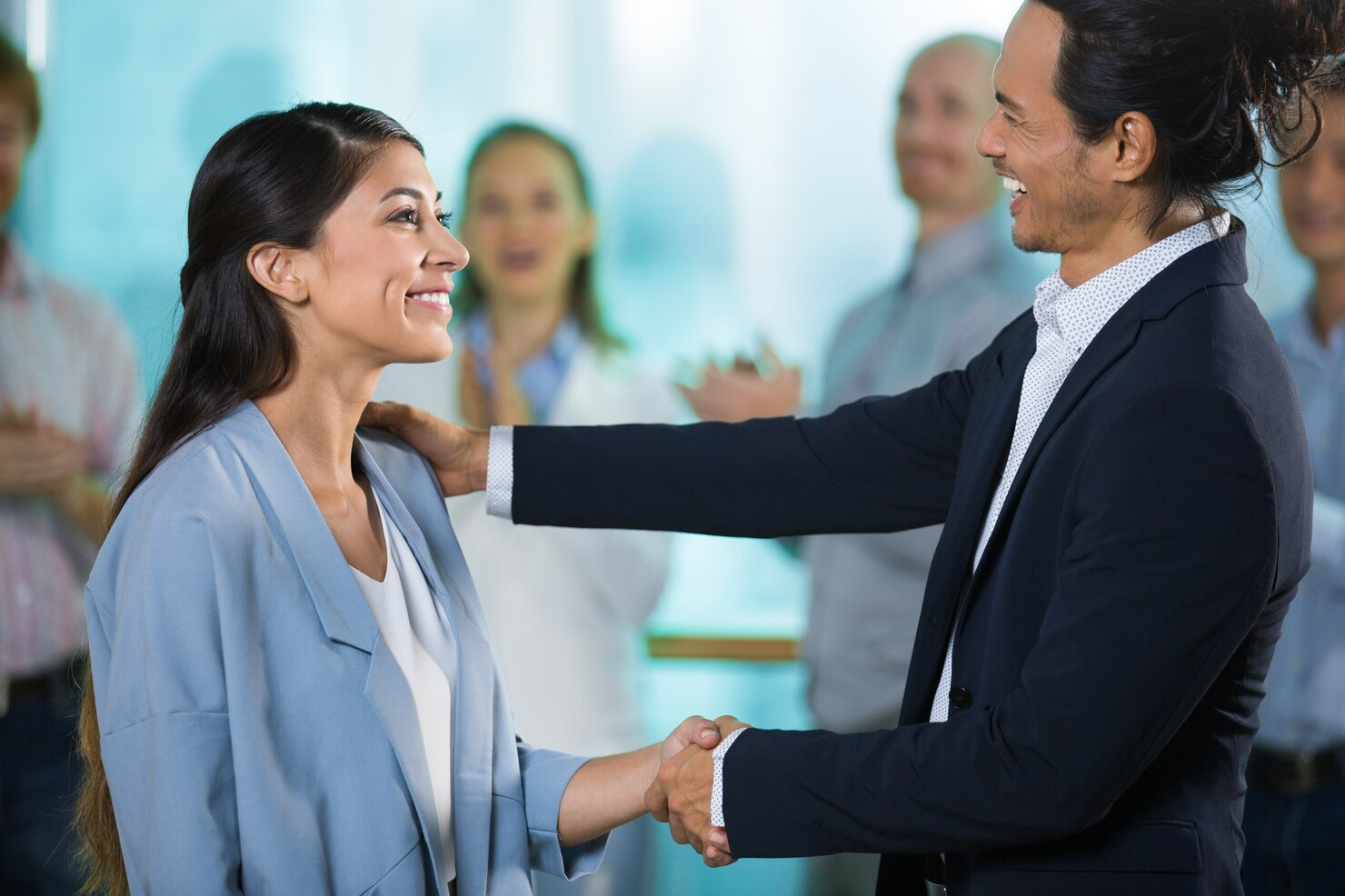 In business attire, a smiling man shakes hands with a smiling woman and pats her shoulder while people clap in the background. 